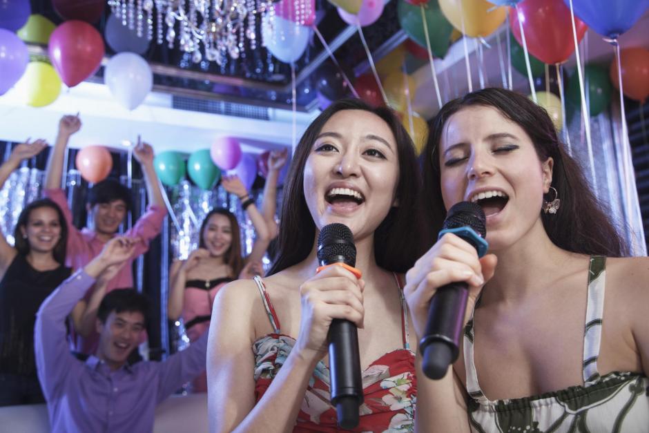 Two friends holding microphones and singing together at karaoke, balloons in the background