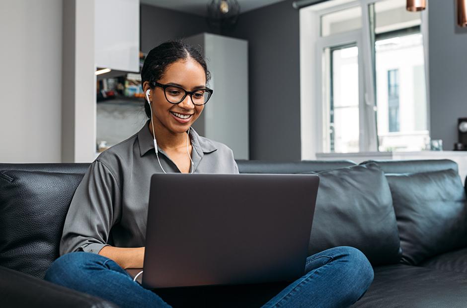 Young woman video calling using a laptop sitting on a sofa wearing earphones 