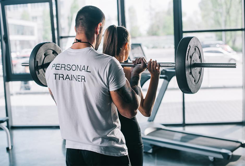 Male personal trainer helping sportswoman to do exercises with barbell at gym