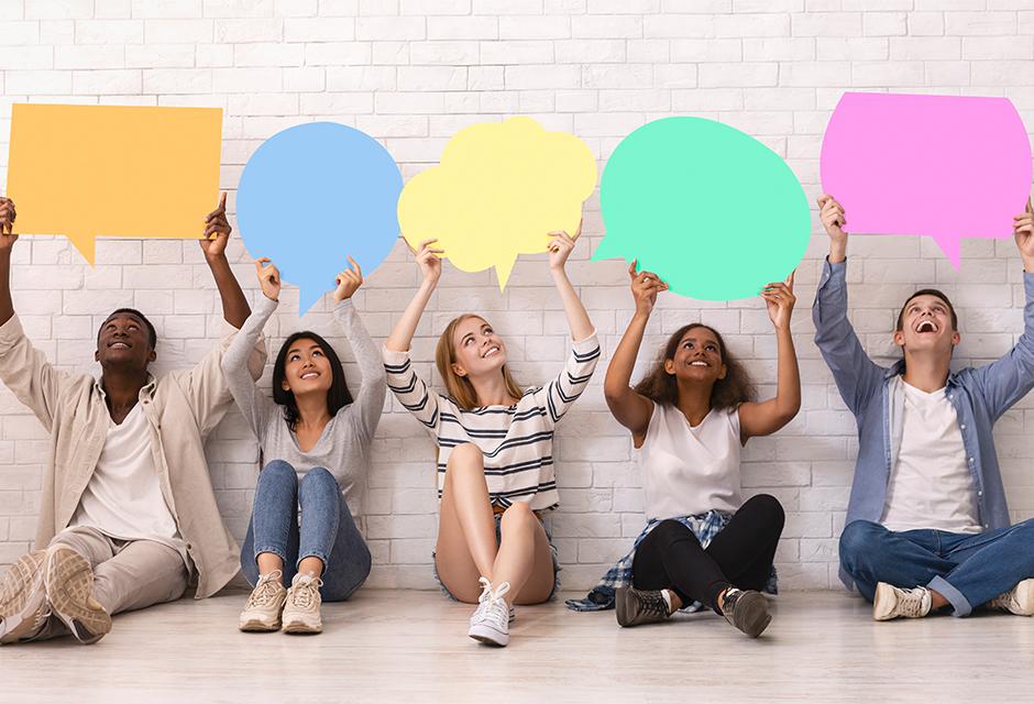 Multiracial group of teenagers sitting on floor and holding colorful speech bubbles above