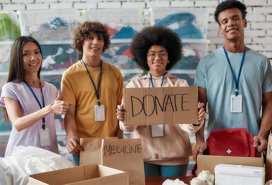 Group of diverse young volunteers smiling at camera while packing medicine donation in paper bags and boxes, Girl holding card with Donate lettering