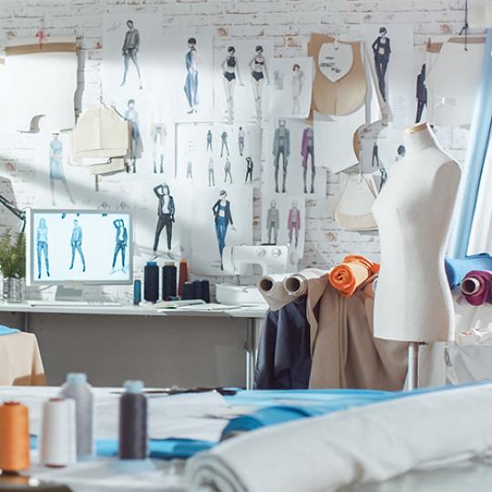 Shot of a Sunny Fashion Design Studio. We See Working Personal Computer, Hanging Clothes, Sewing Machine and Various Sewing Related Items on the Table, Mannequins Standing, Colorful Fabrics.